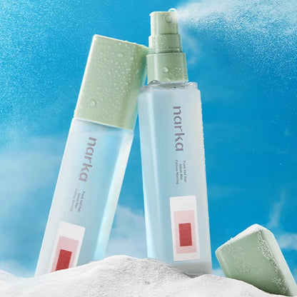 Two Narika skincare bottles and a matching cap placed on white sand against a bright blue sky backdrop with mist and clouds. The bottles are frosted light blue with pale green caps and labels, and a small red square graphic appears near the bottom.